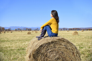 Naklejka na ściany i meble Young brunette woman in yellow sweater and rubber boots sitting on top of haystack in field. she looking at mountains, Japanese sea and the autumn forest. Primorsky kray, Russia. Beauty in nature.