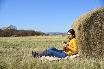 Naklejka na ściany i meble Young Russian brunette woman in yellow sweater and rubber boots sitting on plaid next to backpack, leaning back on bale of hay in field. she holding thermos with tea, trying to open it. Autumn, picnic