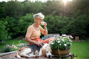 Portrait of senior woman with pet dog sitting outdoors in garden, relaxing with coffee.
