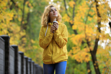 Woman in yellow jacket drinks a tea walking along fall alley