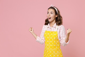 Excited young brunette woman housewife 20s in yellow apron doing winner gesture clenching fists while doing housework isolated on pastel pink colour background studio portrait. Housekeeping concept.