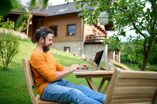 Side view of man with laptop working outdoors in garden, home office concept.