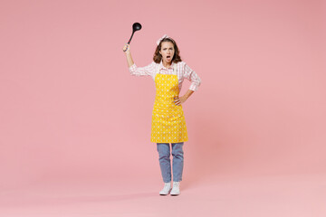 Full length portrait angry displeased young woman housewife in apron hold soup black ladle dipper swearing while doing housework isolated on pastel pink colour background studio. Housekeeping concept.