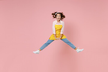 Fototapeta na wymiar Full length portrait of excited young brunette woman housewife 20s in yellow apron jumping spreading legs while doing housework isolated on pastel pink colour background studio. Housekeeping concept.