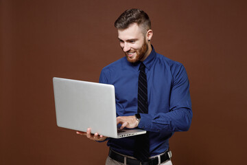 Smiling successful bearded young business man wearing blue shirt tie working on laptop pc computer isolated on brown colour background studio portrait. Achievement career wealth business concept.