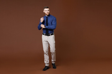 Fototapeta premium Full length of successful bearded young business man in blue shirt tie straightening sleeves looking aside isolated on brown background studio portrait. Achievement career wealth business concept.