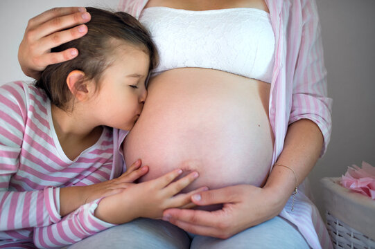 Small girl kissing belly of unrecognizable pregnant mother indoors at home.