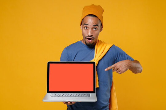 Shocked young african american man in blue t-shirt hat standing pointing index finger on laptop pc computer with blank empty screen mock up copy space isolated on yellow background, studio portrait.