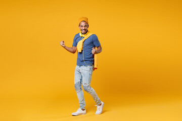 Full length of happy joyful laughing young african american man 20s wearing basic blue t-shirt hat standing clenching fists doing winner gesture isolated on bright yellow background, studio portrait.