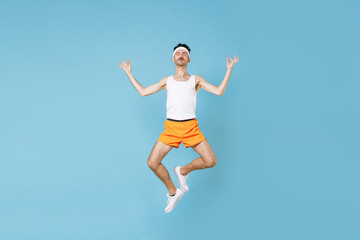 Fototapeta na wymiar Full length young fitness man with skinny body sportsman in headband shirt shorts jumping hold hands in yoga gesture, relaxing meditating isolated on blue background. Workout sport motivation concept.