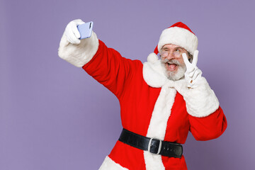 Blinking Santa Claus man in Christmas hat red suit coat glasses doing selfie shot on mobile phone showing victory sign isolated on violet background. Happy New Year celebration merry holiday concept.