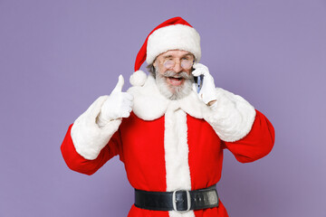 Cheerful Santa Claus man in Christmas hat red suit coat white gloves glasses talking on mobile phone showing thumb up isolated on violet background. Happy New Year celebration merry holiday concept.