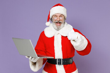 Excited Santa Claus man in Christmas hat red suit coat gloves glasses working on laptop pc computer showing thumb up isolated on violet background. Happy New Year celebration merry holiday concept.