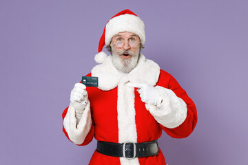 Fototapeta na wymiar Amazed elderly Santa Claus man in Christmas hat red suit coat gloves glasses pointing index finger on credit bank card isolated on violet background. Happy New Year celebration merry holiday concept.