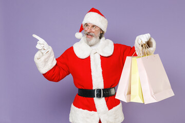 Funny Santa Claus man in Christmas hat suit glasses hold package bags purchases after shopping point index finger aside isolated on violet background. Happy New Year celebration merry holiday concept.
