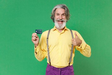 Excited elderly gray-haired mustache bearded man in casual yellow shirt suspenders hold credit bank card showing thumb up looking camera isolated on bright green colour background studio portrait.