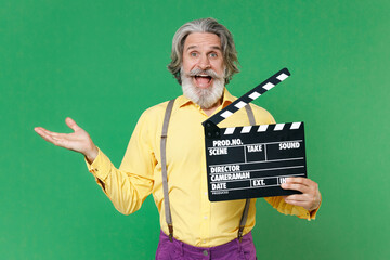 Excited elderly gray-haired mustache bearded man wearing basic yellow shirt suspenders hold classic black film making clapperboard spreading hands isolated on green colour background, studio portrait.