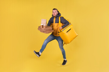 Blond unshaven teen food delivery boy and courier in yellow clothes with thermo bag on his back holds an order for customer and runs to deliver order on time in 15 minutes. Food delivery in city