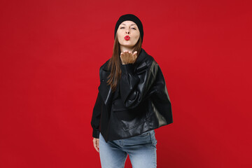 Beautiful attractive pretty young brunette woman wearing casual black leather jacket white t-shirt hat blowing sending air kiss looking camera isolated on bright red colour background studio portrait.