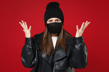 Shocked worried young woman 20s wearing black leather jacket hat face mask safe from coronavirus virus covid-19 during pandemic quarantine spreading hands isolated on red background studio portrait.