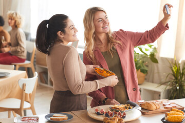 Portrait of two modern adult women taking selfie photo indoors while enjoying dinner party with friends, copy space