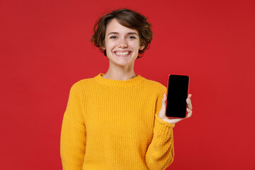 Smiling young brunette woman 20s wearing casual yellow sweater standing hold mobile cell phone with blank empty screen mock up copy space isolated on bright red colour background studio portrait.