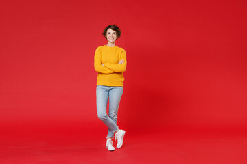 Full length of cheerful attractive young brunette woman 20s wearing basic casual yellow sweater standing holding hands crossed looking camera isolated on bright red colour background studio portrait. - obrazy, fototapety, plakaty
