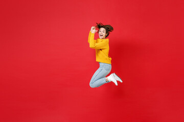 Full length side view of excited cheerful joyful young brunette woman 20s in basic yellow sweater jumping clenching fists doing winner gesture isolated on bright red colour background studio portrait.