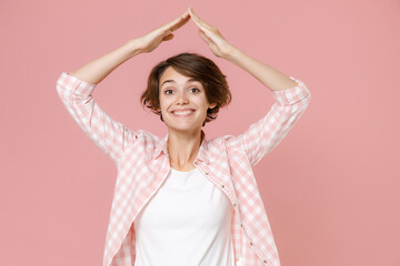 Smiling cheerful young brunette woman 20s wearing casual checkered shirt standing holding hands above head like roof of house looking camera isolated on pastel pink colour background, studio portrait.