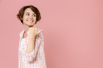 Obraz premium Side view of smiling pretty young brunette woman 20s wearing casual checkered shirt standing pointing thumb aside on mock up copy space isolated on pastel pink colour background, studio portrait.
