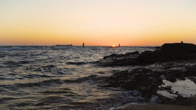 El agua del mar con olas en una orilla rocosa y la puesta de sol con una isla con un castillo al fondo. desde la playa de la barrosa, en Chiclana, C&aacute;diz, Andaluc&iacute;a, sur de Espa&ntilde;a.