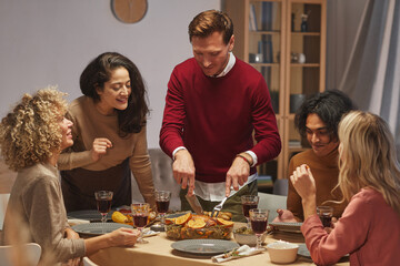 Portrait of smiling adult man cutting delicious roasted turkey while enjoying Thanksgiving dinner with friends and family, copy space