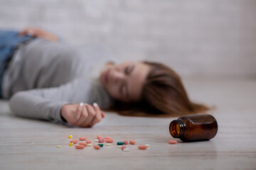 Young woman lying on floor with bottle of tablets scattered on floor, committing suicide, selective focus