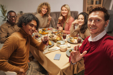 Fototapeta na wymiar Portrait of smiling adult man looking at camera while taking selfie photo with friends and family at Thanksgiving dinner, copy space