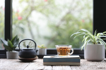 Transparent cup tea with books and black tea pot and plants on wooden table in front of windows
