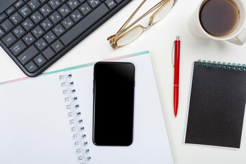 a phone with a screen mockup, a coffee cup of writing supplies, a pens, a notepad on a white wooden table background. Top view of the work area, copy space