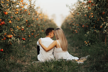 Happy couple together are relaxing and drinking wine at summer picnic in the apples orchard. Back view.