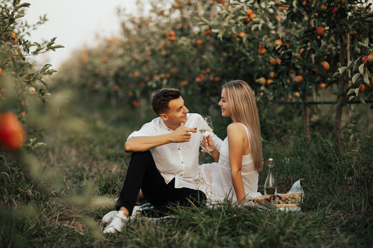 Happy couple in love have a picnic in the summer apple orchard. They clink glasses with white wine.