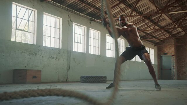 Fitness man working out with battle ropes at cross training gym. Athletic male model with battle rope doing exercise inside empty warehouse.
