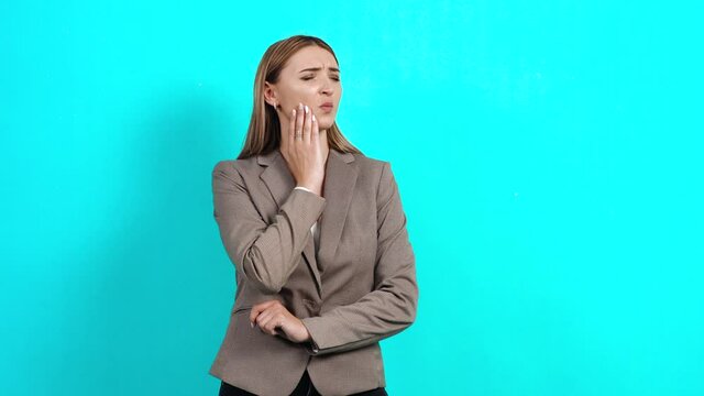Frustrated young woman in business suit, touching her cheek, feeling sharp toothache, upset with gum disease, suffering from tartar, dentistry. Studio image indoors, isolated on blue background