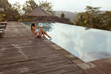 Beautiful Girl In Bikini Sitting Near Infinity Pool In Bali, Indonesia. Young Woman’s With Sexy Body In White Swimsuit Relaxing At Tropical Resort Portrait.