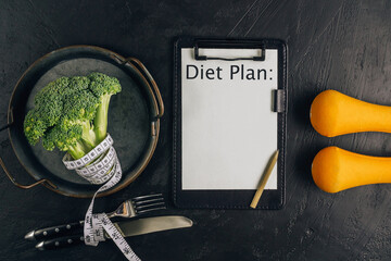 Top view of Diet plan on paper with broccoli, cutlery, measuring tape and dumbbells on black table.