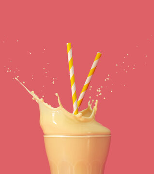 drinking straws into a splashing glass of yellow milkshake on pastel pink background.