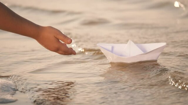 Hand of a little girl lowers a paper boat into the river. Children's paper boat on the waves of the river. Close-up of a little girl's hand with a paper boat