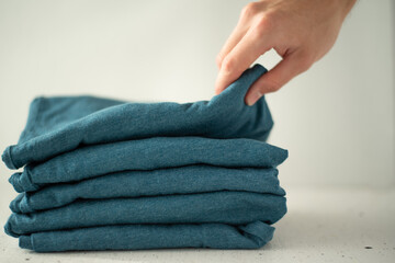 A stack of t-shirts of the same color on a light background. A man's hand picks up the top t-shirt from the stack. The minimalistic concept. Space for text.