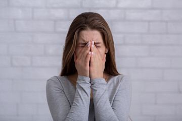 Depressed millennial woman covering her face and crying against white brick wall