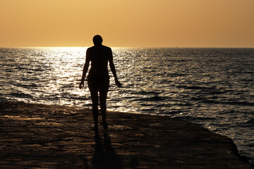 Fototapeta premium Silhouette of woman in bikini walking by the rocky beach on sunset and dark sea background. Romantic vacation and swimming concept