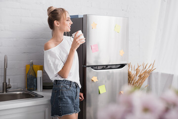selective focus of blonde woman in white t-shirt and denim shorts standing in kitchen with cup of...
