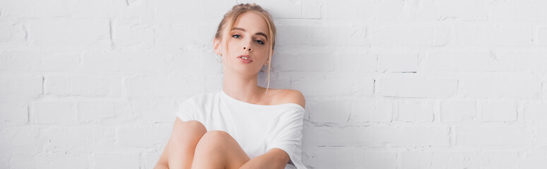 panoramic orientation of young blonde woman looking at camera while sitting near white wall - obrazy, fototapety, plakaty