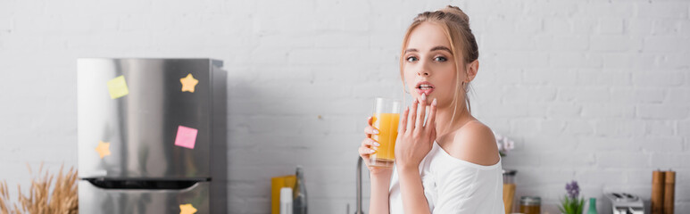 horizontal concept of blonde woman holding glass of orange juice and touching lips in kitchen